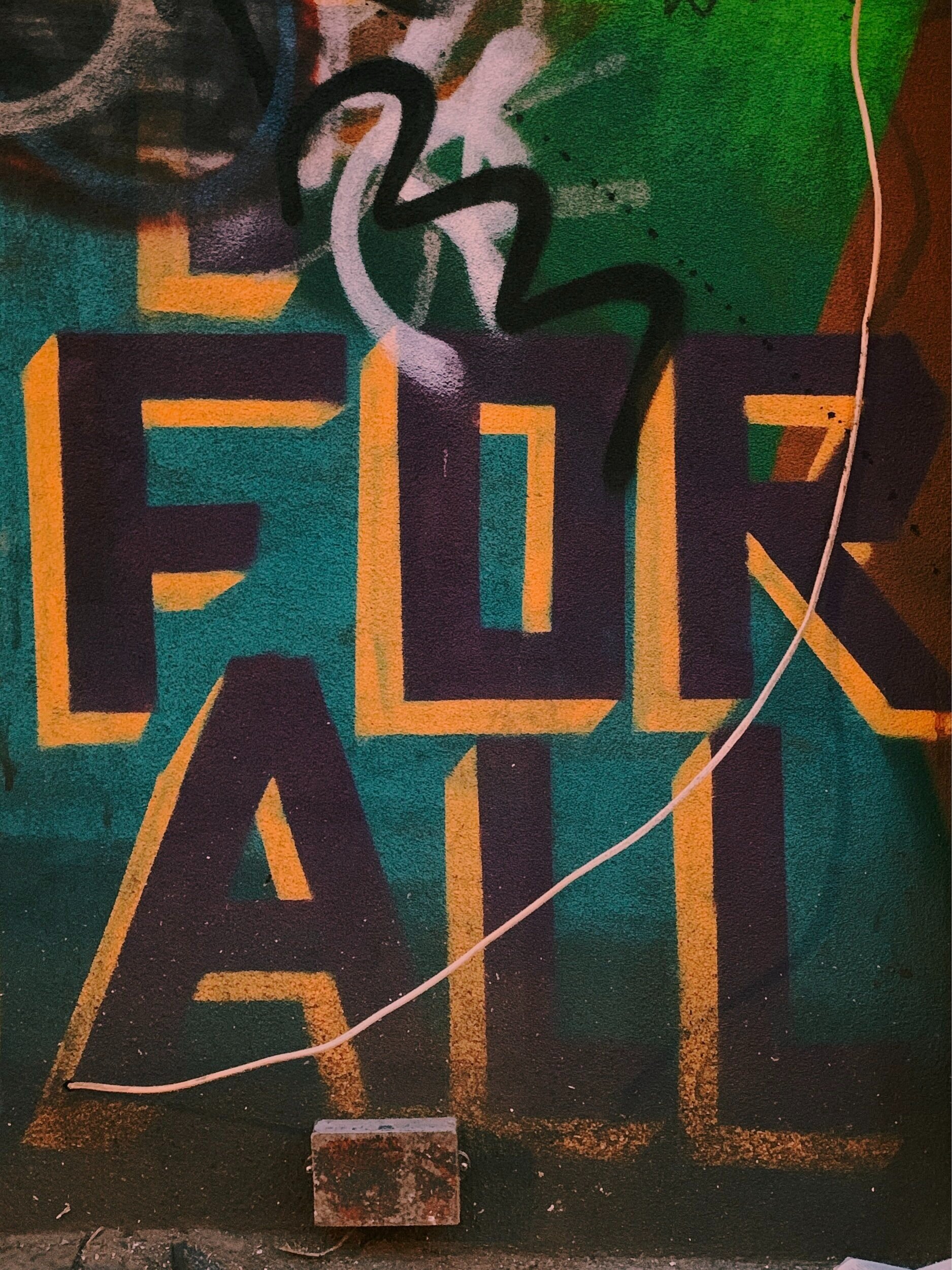 Graffiti on a wall displaying the words "FOR ALL" in large, bold letters. The background features green and orange tones with abstract shapes and a cable hanging loosely across the image.