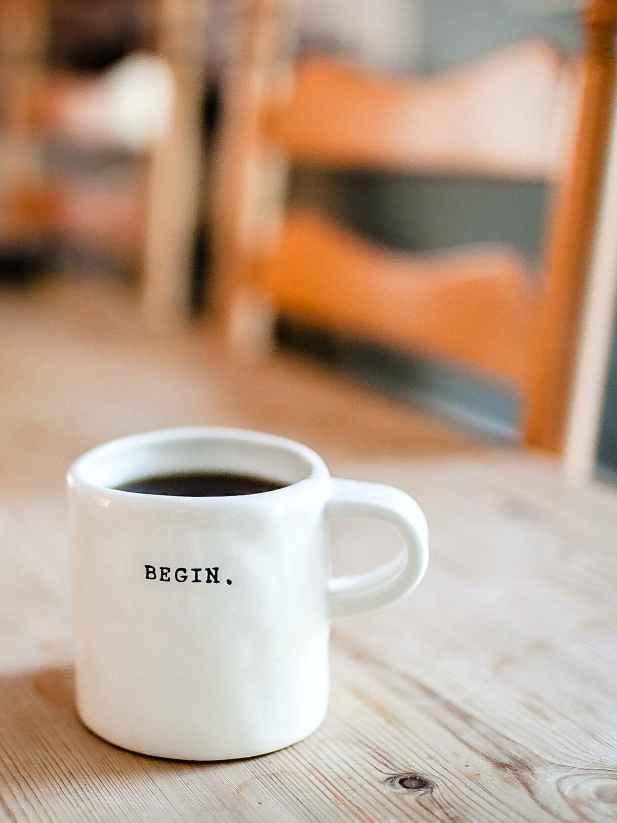A white mug filled with coffee sits on a light wooden table. The mug has the word "BEGIN." printed in black on its side. In the background, there are blurred wooden chairs.