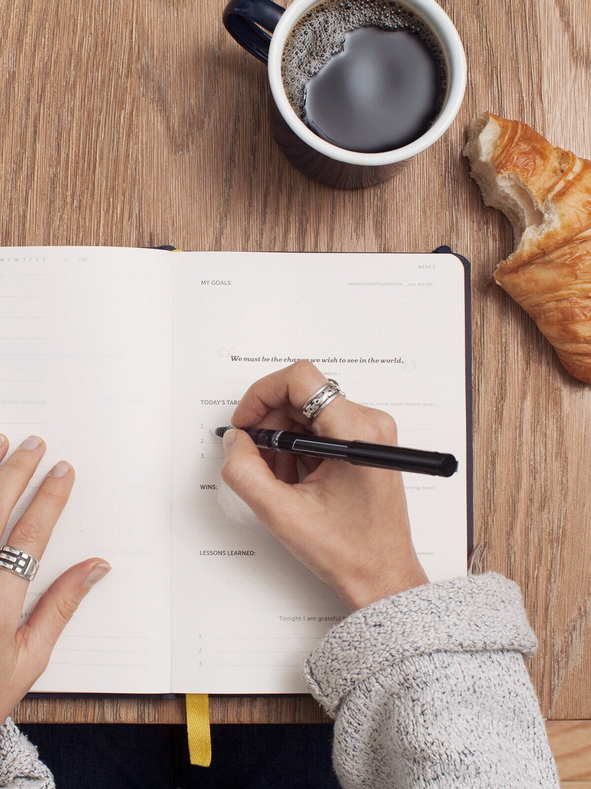 A person wearing a gray sweater writes in a journal on a wooden table. Nearby are a cup of coffee and a partially eaten croissant. The journal has sections for goals, tasks, and lessons learned.