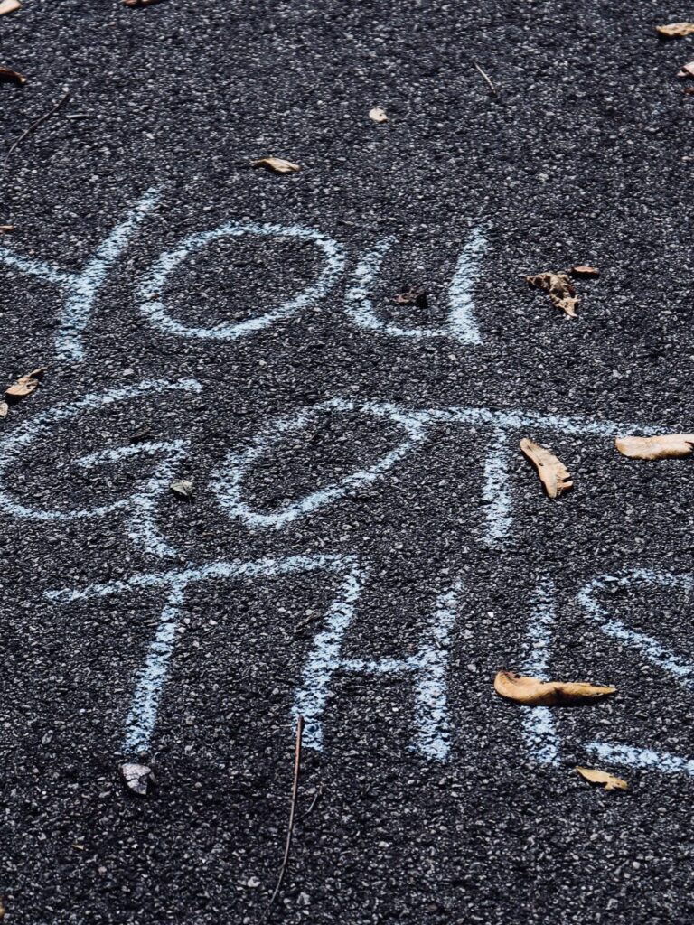 Chalk writing on pavement reads "You Got This" with scattered fallen leaves around it.