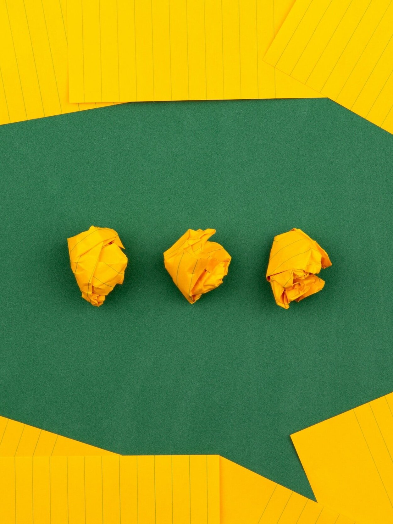 Three crumpled yellow paper balls are placed in a row on a green background shaped like a speech bubble. Lined, flat yellow sheets surround the bubble, creating a colorful contrast.