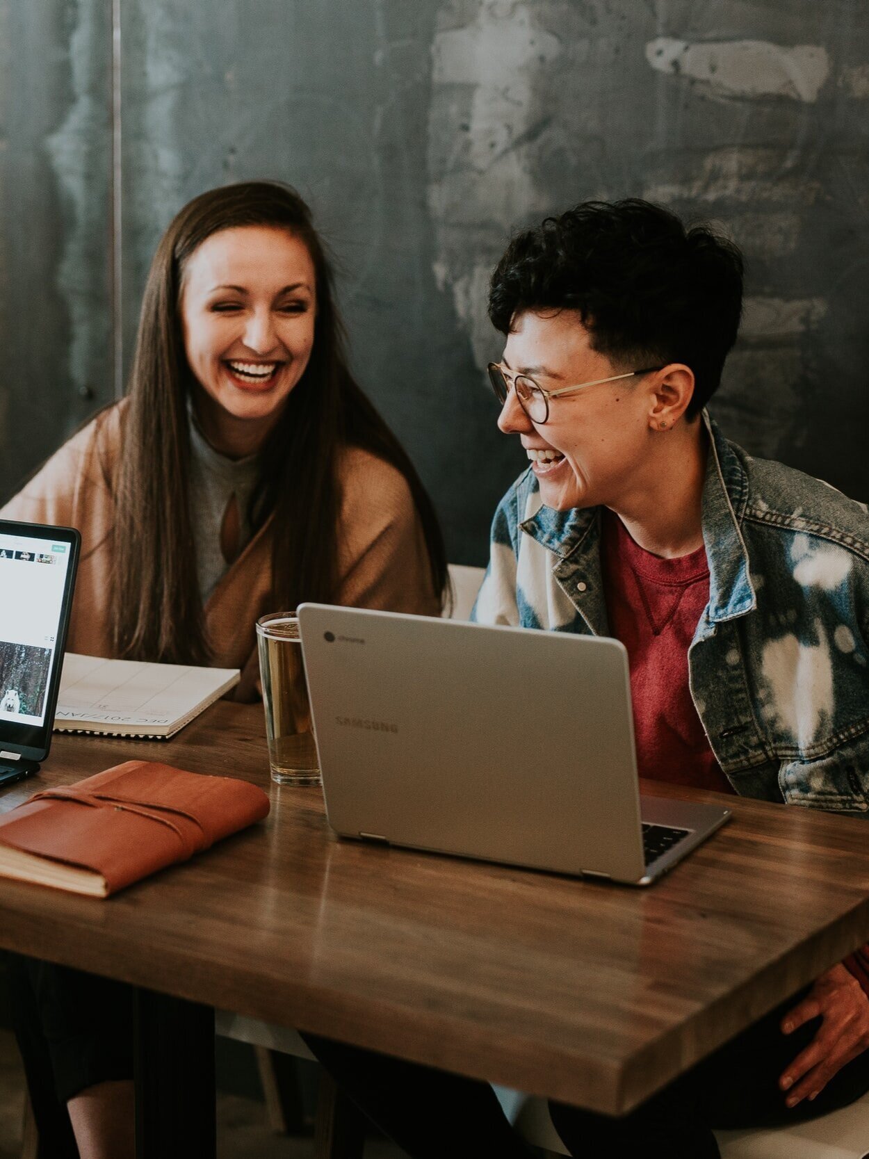 Two people are sitting at a wooden table with laptops, smiling and laughing. One has long hair, the other short hair and glasses. A notebook and a glass sit on the table. A chalkboard wall is in the background.