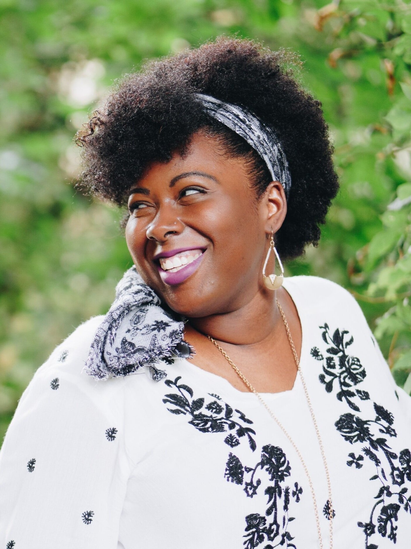 A person with curly hair and a headband smiles while looking to the side. They are wearing a white top with black floral embroidery and geometric earrings. The background is filled with greenery.