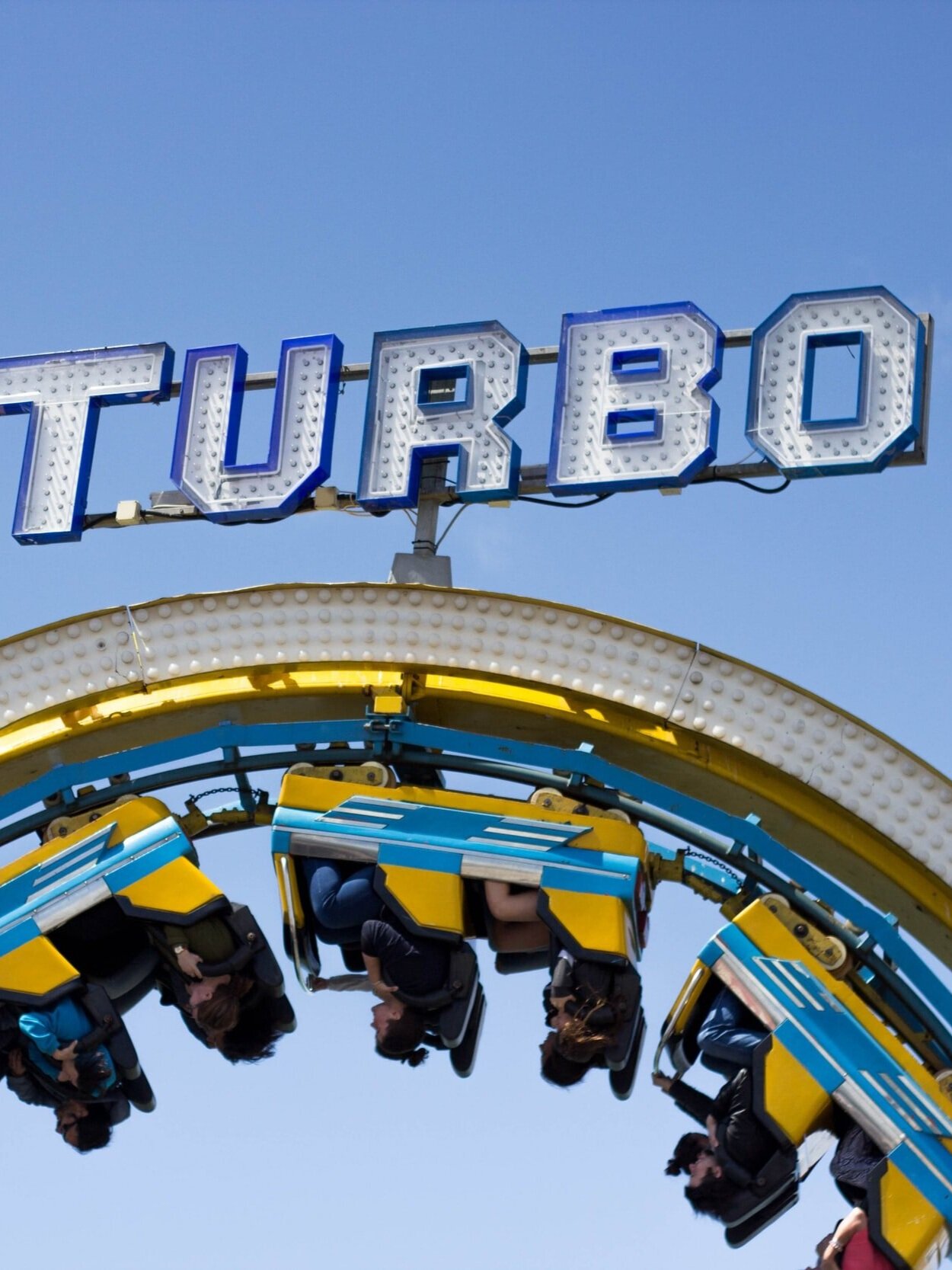 A roller coaster with people riding in blue and yellow cars is upside down on a loop against a clear blue sky. The large, illuminated sign above reads "TURBO.