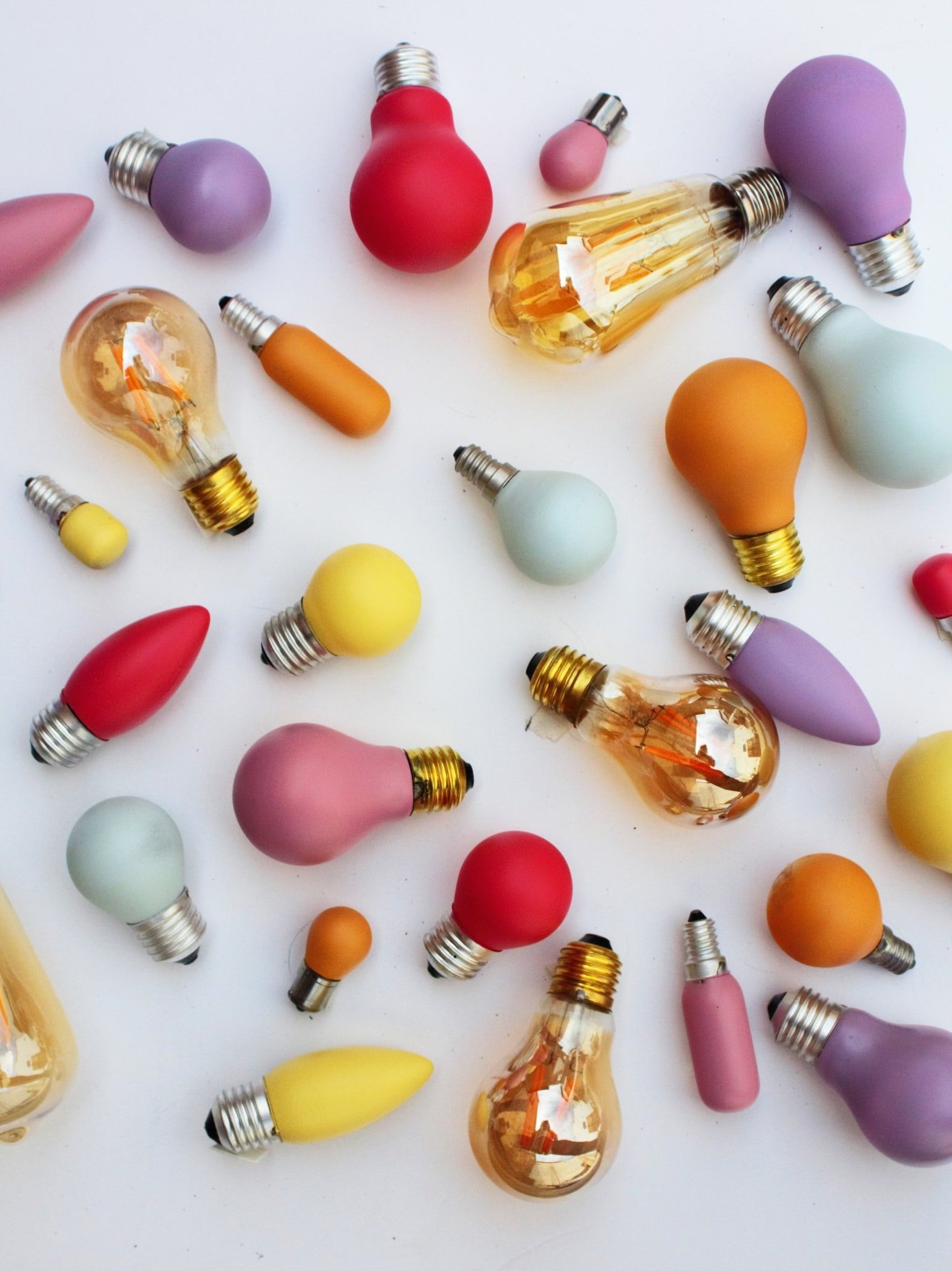 A collection of colorful light bulbs scattered on a white background. The bulbs vary in shape and color, including shades of red, yellow, purple, orange, and clear glass, showcasing an artistic arrangement.