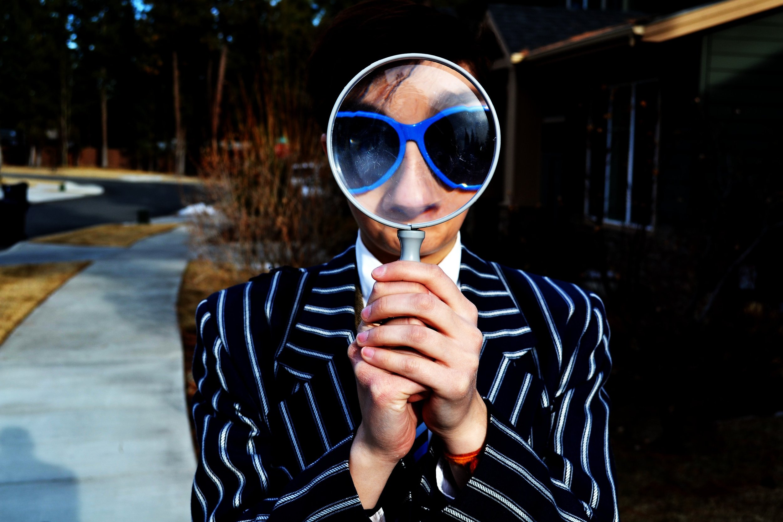 A person wearing a black and white striped jacket looks through a magnifying glass, which enlarges their blue sunglasses. They are standing outside on a path with trees and buildings in the background.