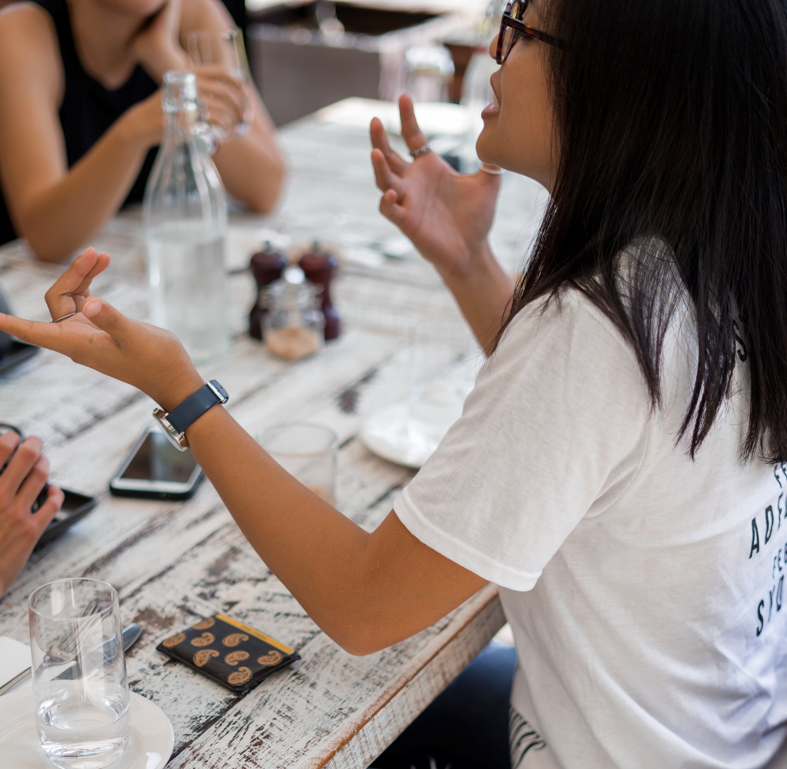 A woman wearing glasses and a white t-shirt is animatedly talking at a wooden table. There are empty glasses, a water bottle, and a phone on the table. Two other people are partially visible, engaged in the conversation.