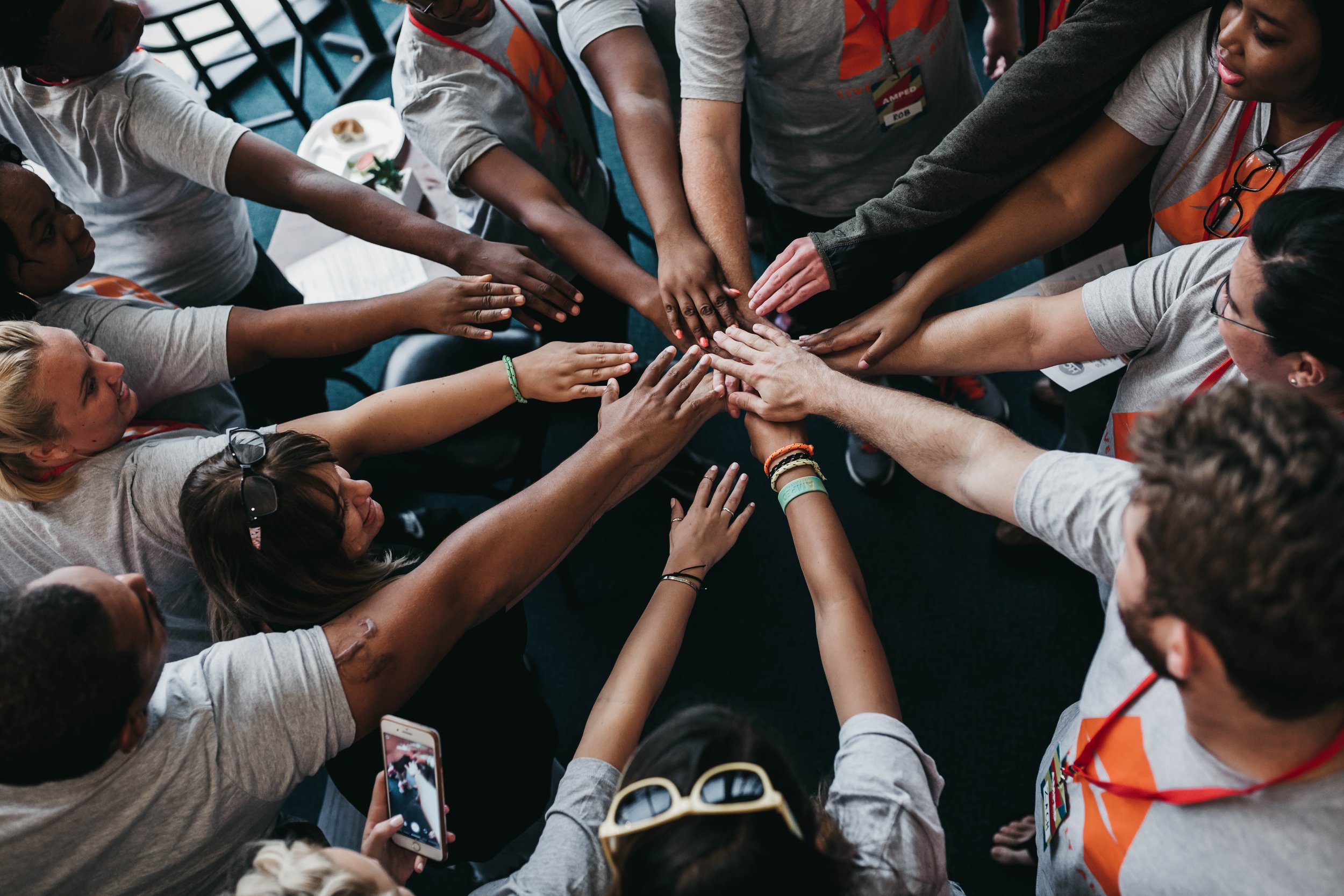 A diverse group of people in gray shirts are standing in a circle, each with their hand extended into the center, showing unity and teamwork. Some wear ID lanyards, and one person is holding a phone in their other hand.