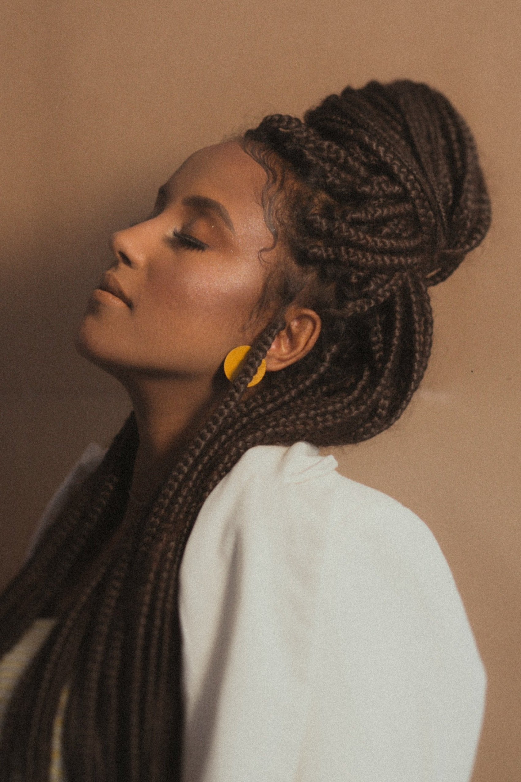 Side profile of a person with eyes closed, wearing a white garment. Their hair is styled in long, thick braids gathered into a high bun. They are wearing yellow earrings and the background is a warm, brown tone.