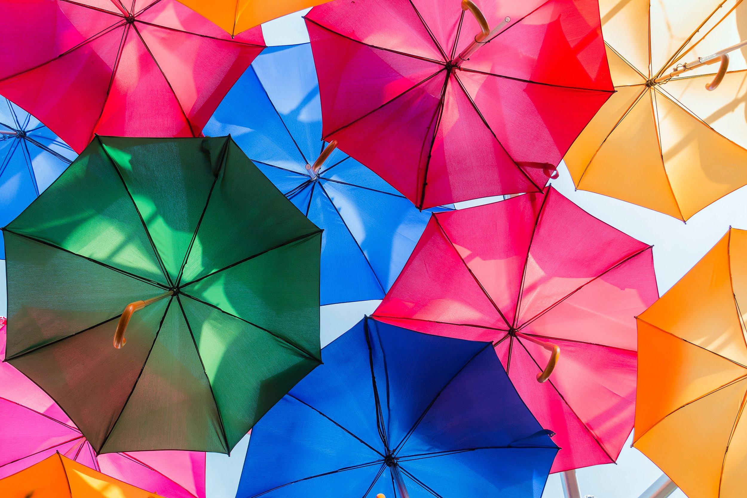 A vibrant display of colorful umbrellas, including green, blue, pink, red, orange, and yellow, viewed from below against a clear blue sky.