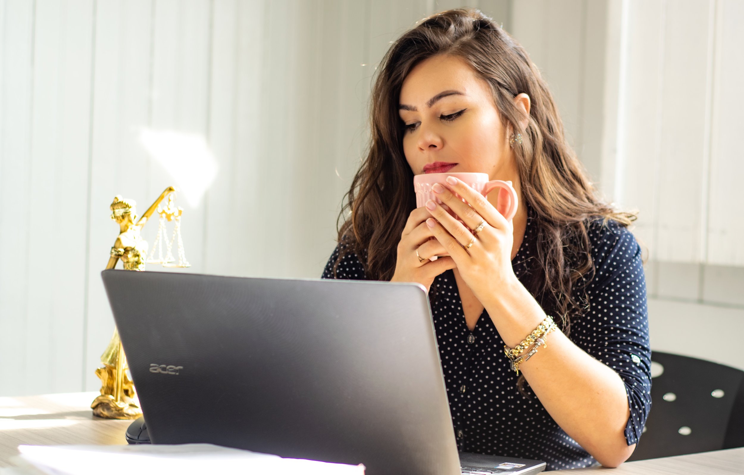 Woman with long hair in a dark polka dot shirt sits at a desk, holding a pink mug, in front of a laptop. A golden lady justice statue is beside her. The room is well-lit with a large window in the background.