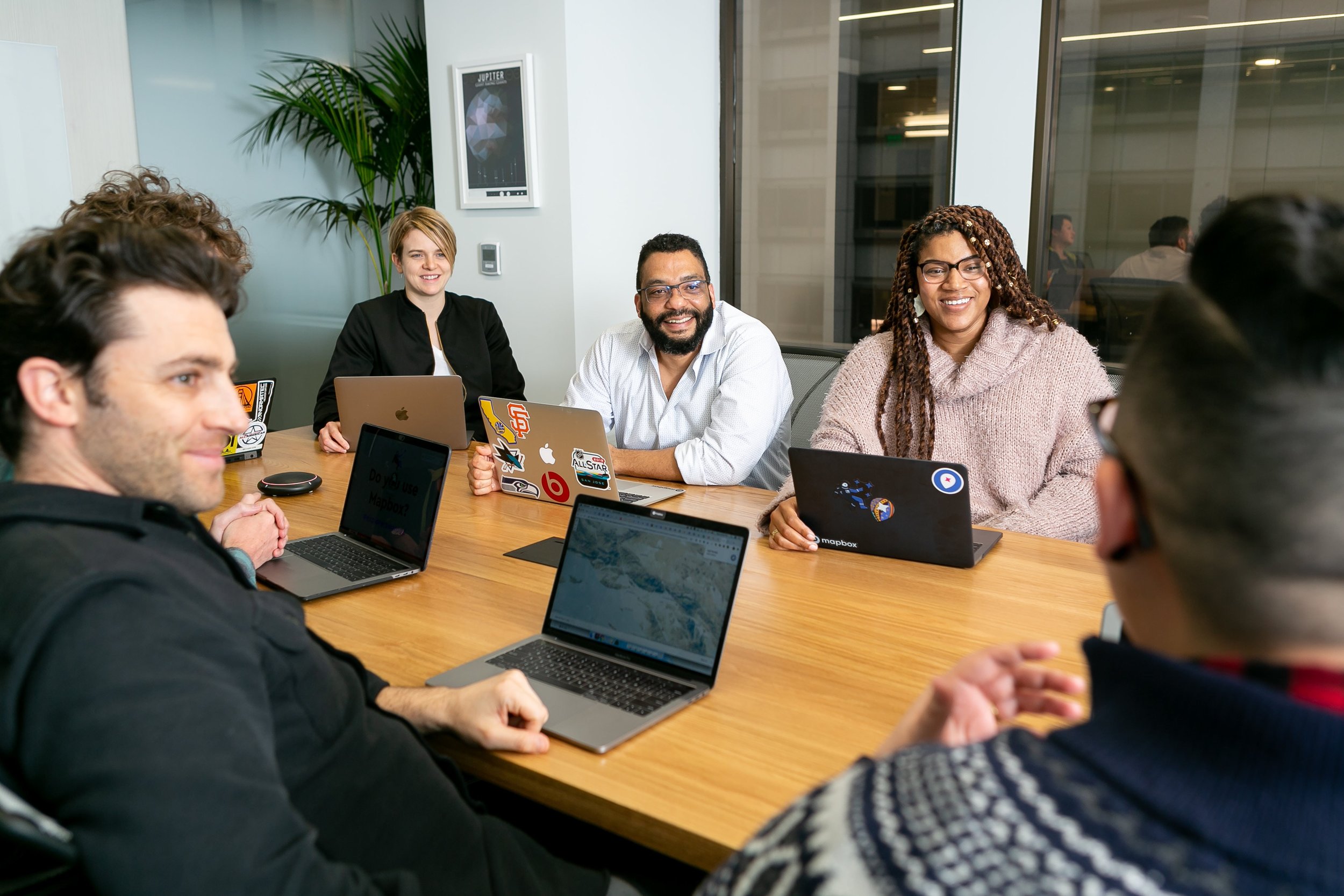 A diverse group of people are sitting around a conference table with laptops open, smiling and engaging in conversation. A plant and a cityscape are visible in the background through the window.