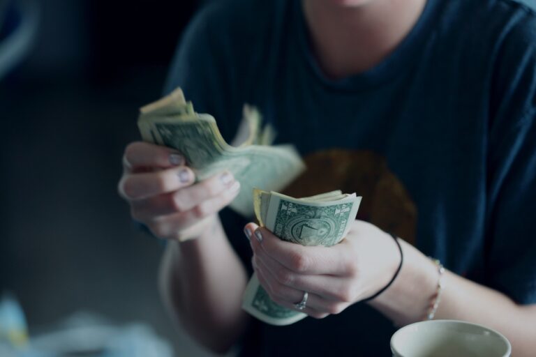 A person wearing a dark blue shirt is counting a stack of US dollar bills. The focus is on their hands and the money, with a blurred background. A white cup is partially visible in the lower right corner.