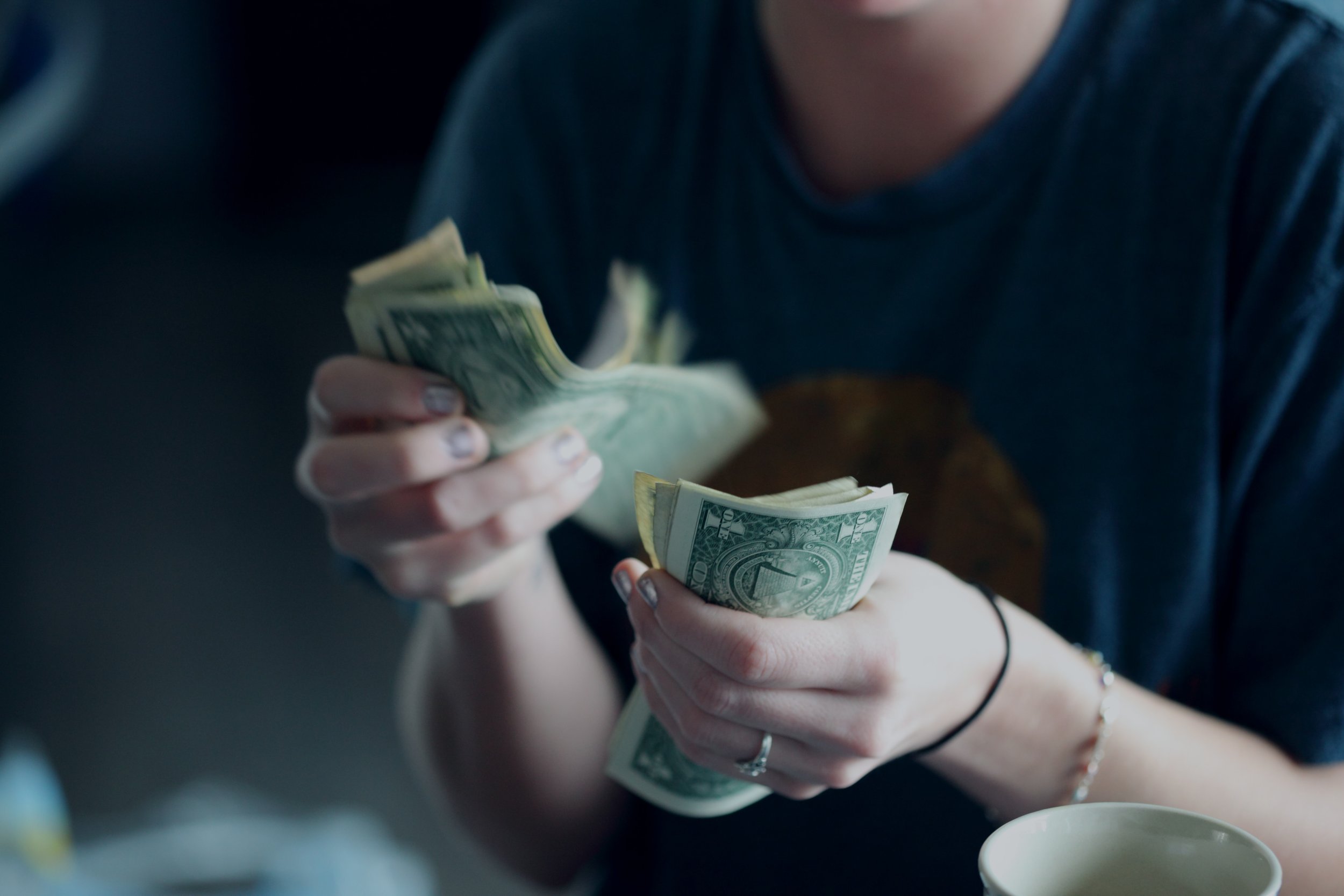 A person wearing a dark blue shirt is counting a stack of US dollar bills. The focus is on their hands and the money, with a blurred background. A white cup is partially visible in the lower right corner.