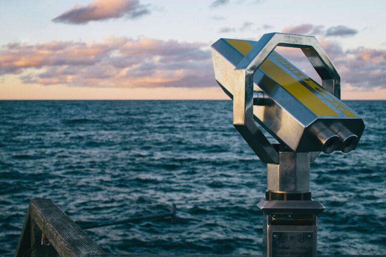 A silver binocular viewer on a wooden platform overlooks a vast, calm ocean under a sky with scattered clouds at sunset, casting a warm glow over the scene.