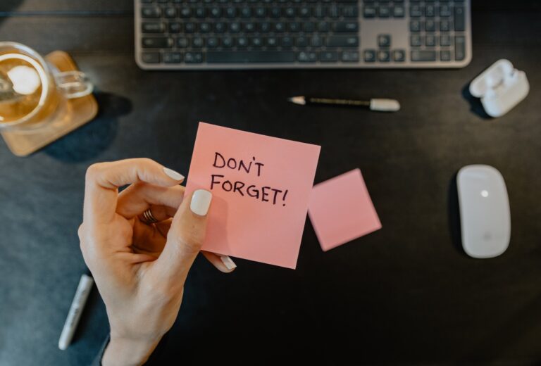 A hand holding a pink sticky note with the message "Don't Forget!" written on it. The background shows a dark desk with a keyboard, a pencil, a mouse, a cup, and another sticky note.