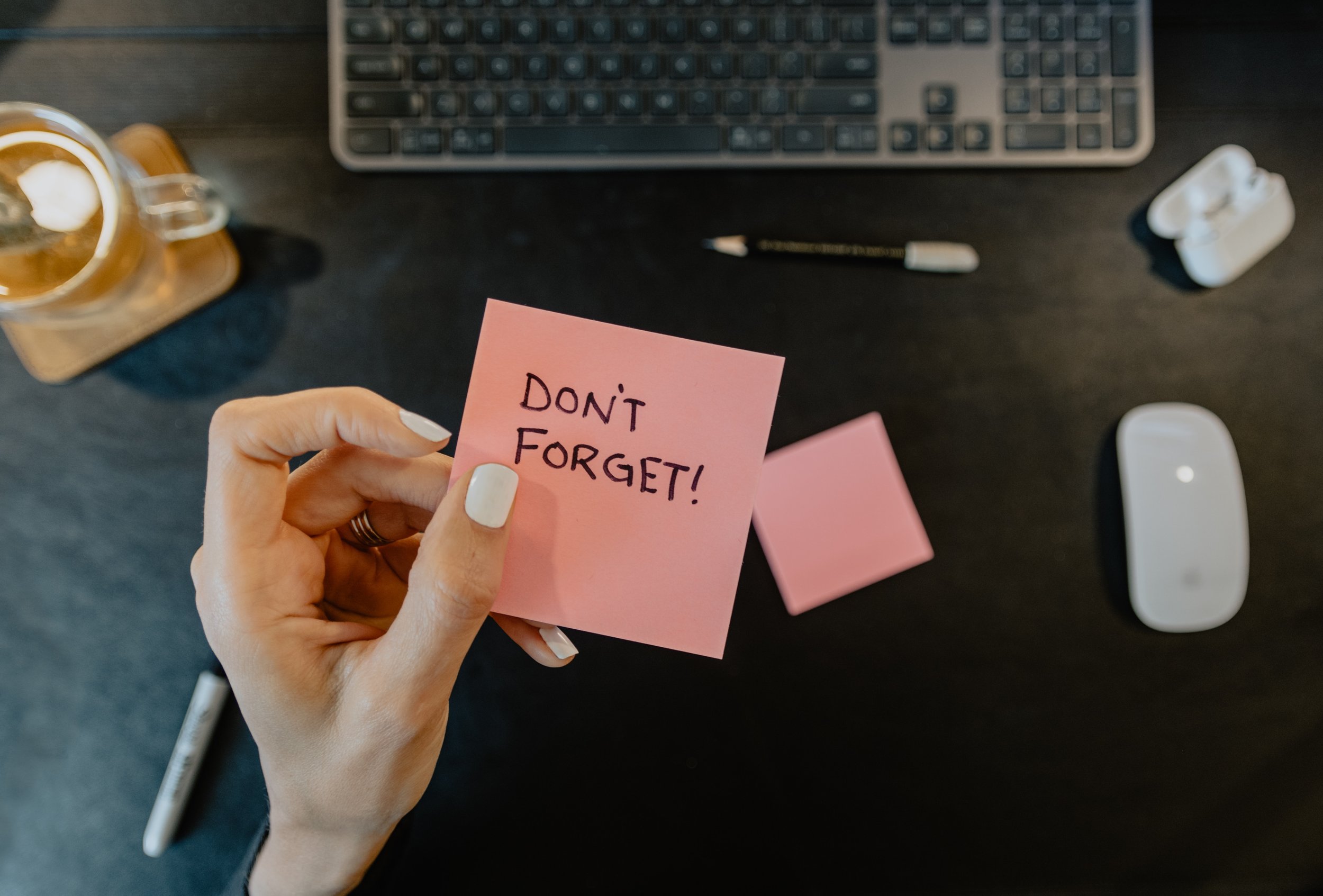 A hand holding a pink sticky note with the message "Don't Forget!" written on it. The background shows a dark desk with a keyboard, a pencil, a mouse, a cup, and another sticky note.