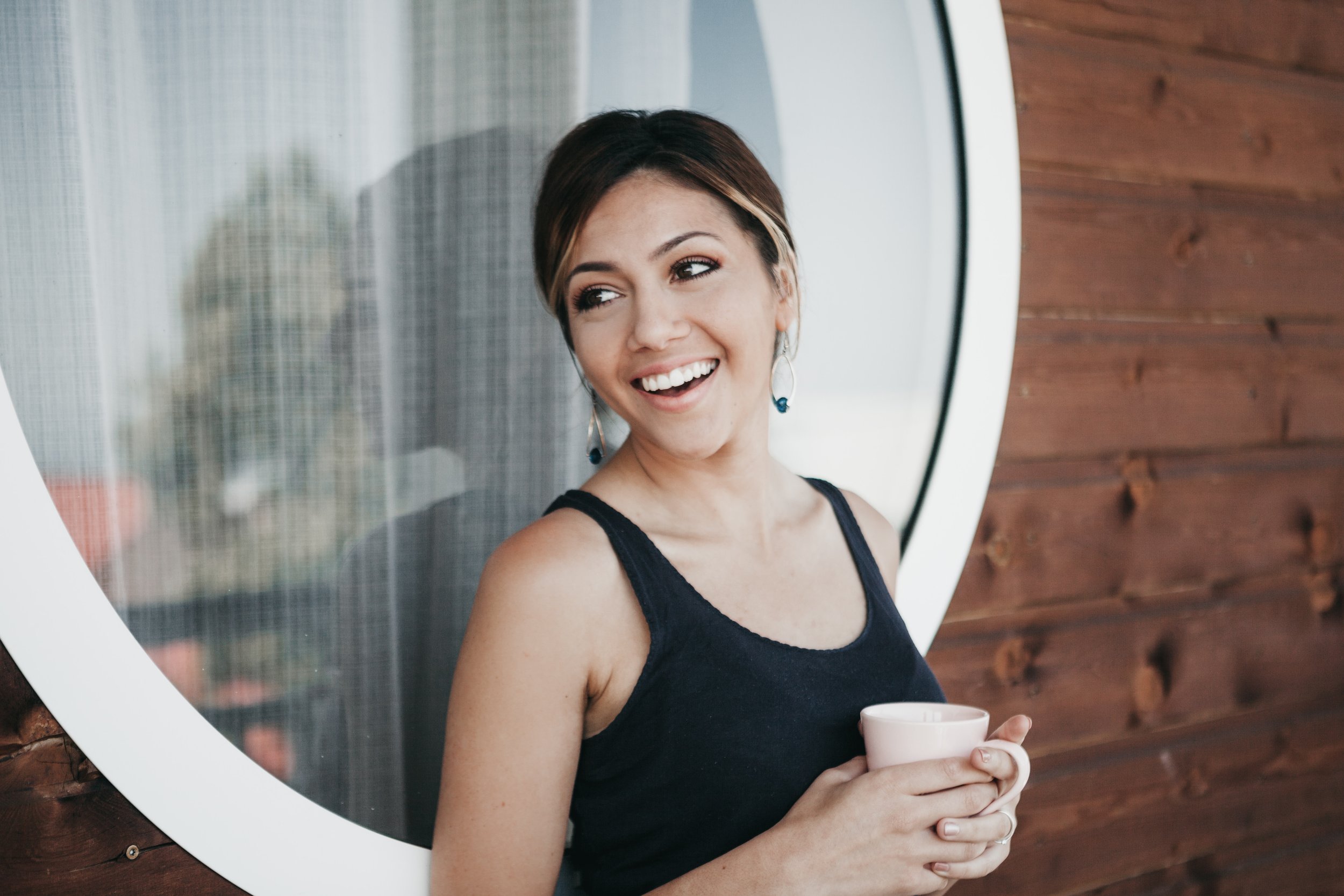 A smiling woman with a light pink mug leans against a round window on a wooden wall. She wears a black tank top and her hair is styled in an updo. The background is slightly blurred, featuring a curtain and hints of greenery.