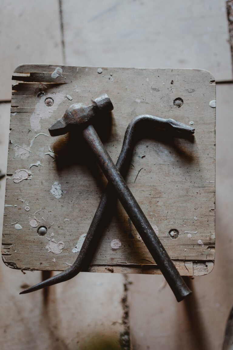 A weathered hammer and crowbar are crossed on a rustic wooden stool. The wood surface is splattered with paint and shows marks of wear, suggesting frequent use. The background features light-colored tiles.