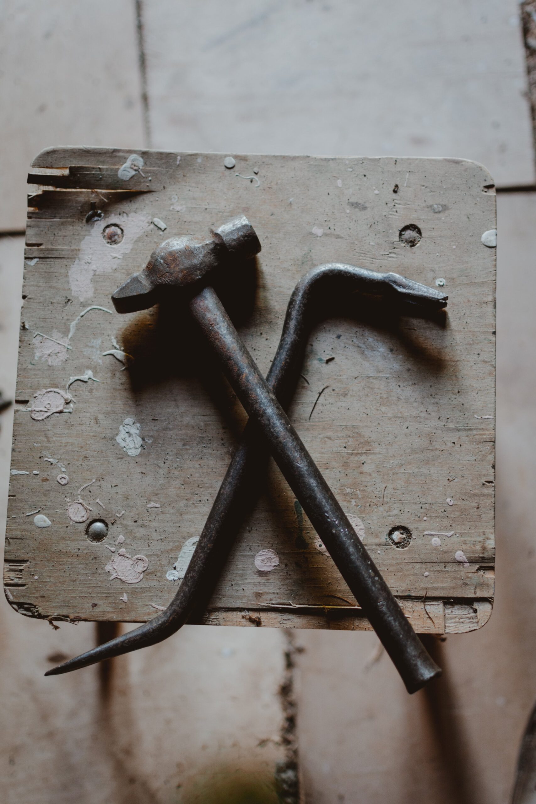 A weathered hammer and crowbar are crossed on a rustic wooden stool. The wood surface is splattered with paint and shows marks of wear, suggesting frequent use. The background features light-colored tiles.