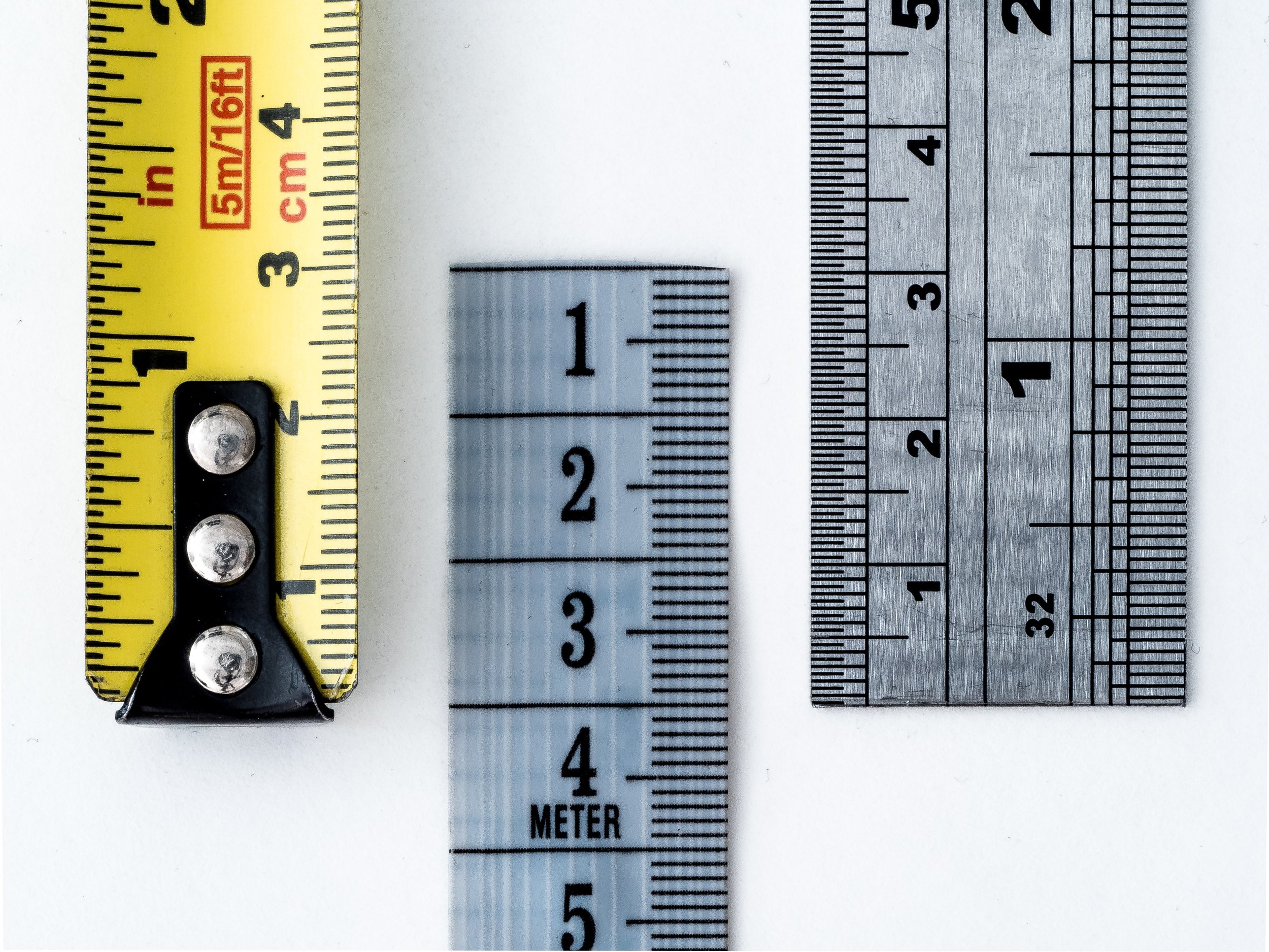 Three measuring tools are displayed vertically against a white background: a yellow tape measure on the left, a clear plastic ruler in the center, and a silver metal ruler on the right.