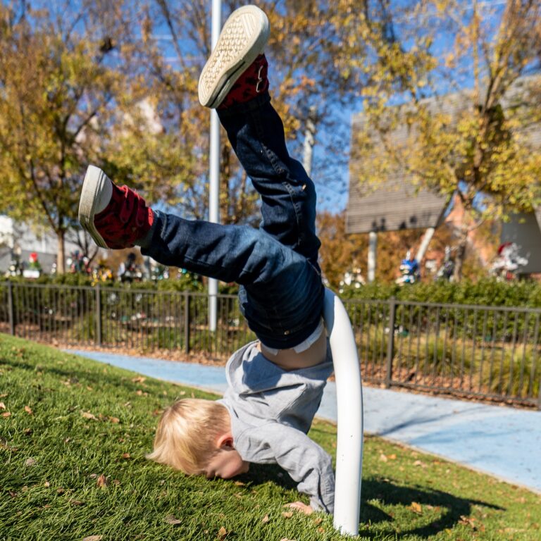 A young child in a gray shirt and jeans is doing a handstand on a grassy slope, with legs bent and feet in the air. The background shows autumn trees and a playground.