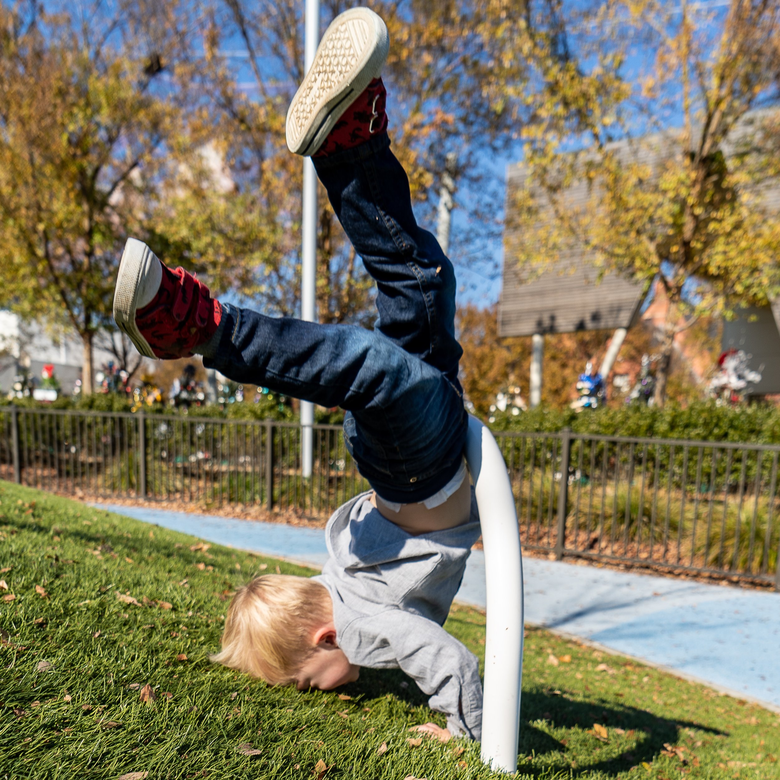 A young child in a gray shirt and jeans is doing a handstand on a grassy slope, with legs bent and feet in the air. The background shows autumn trees and a playground.