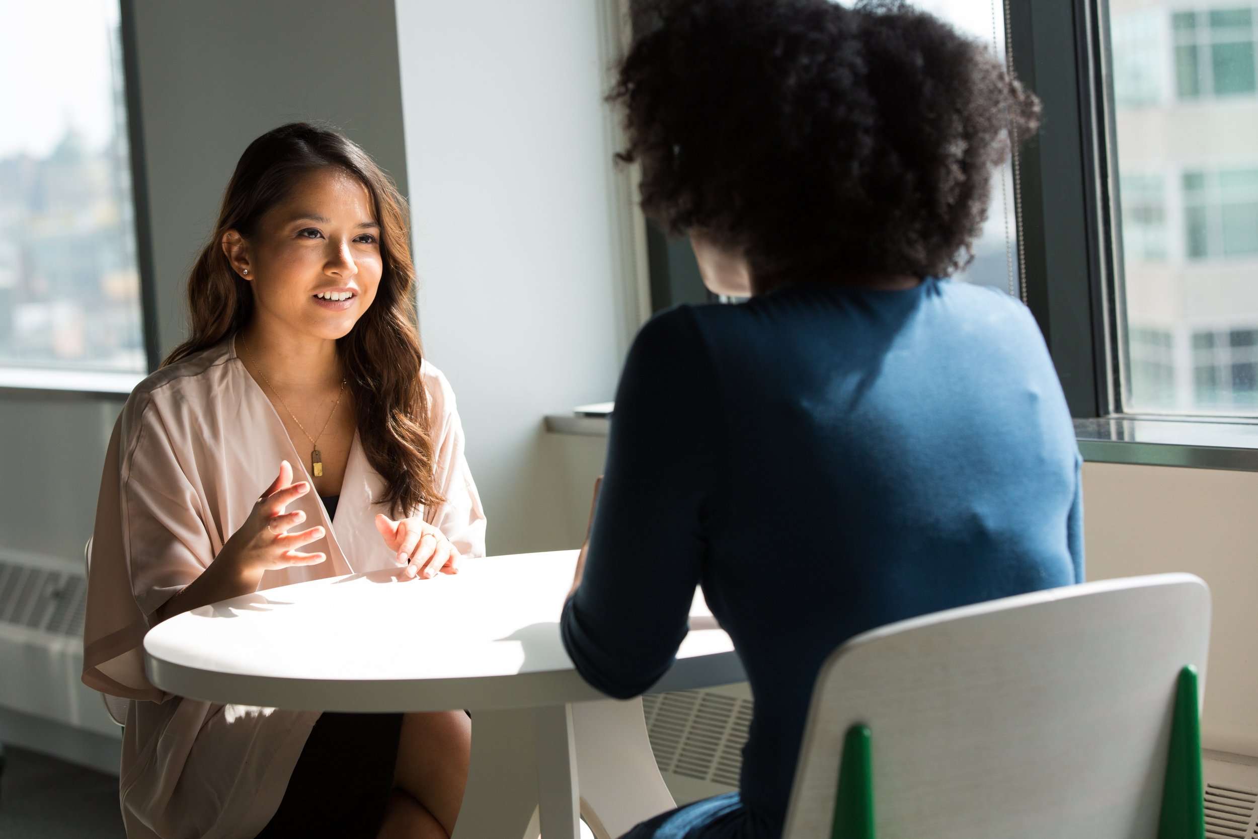 Two women are sitting at a round table in a modern office setting. One woman, facing the camera, is gesturing while speaking. The other woman has her back to the camera. They're engaged in a discussion near large windows.
