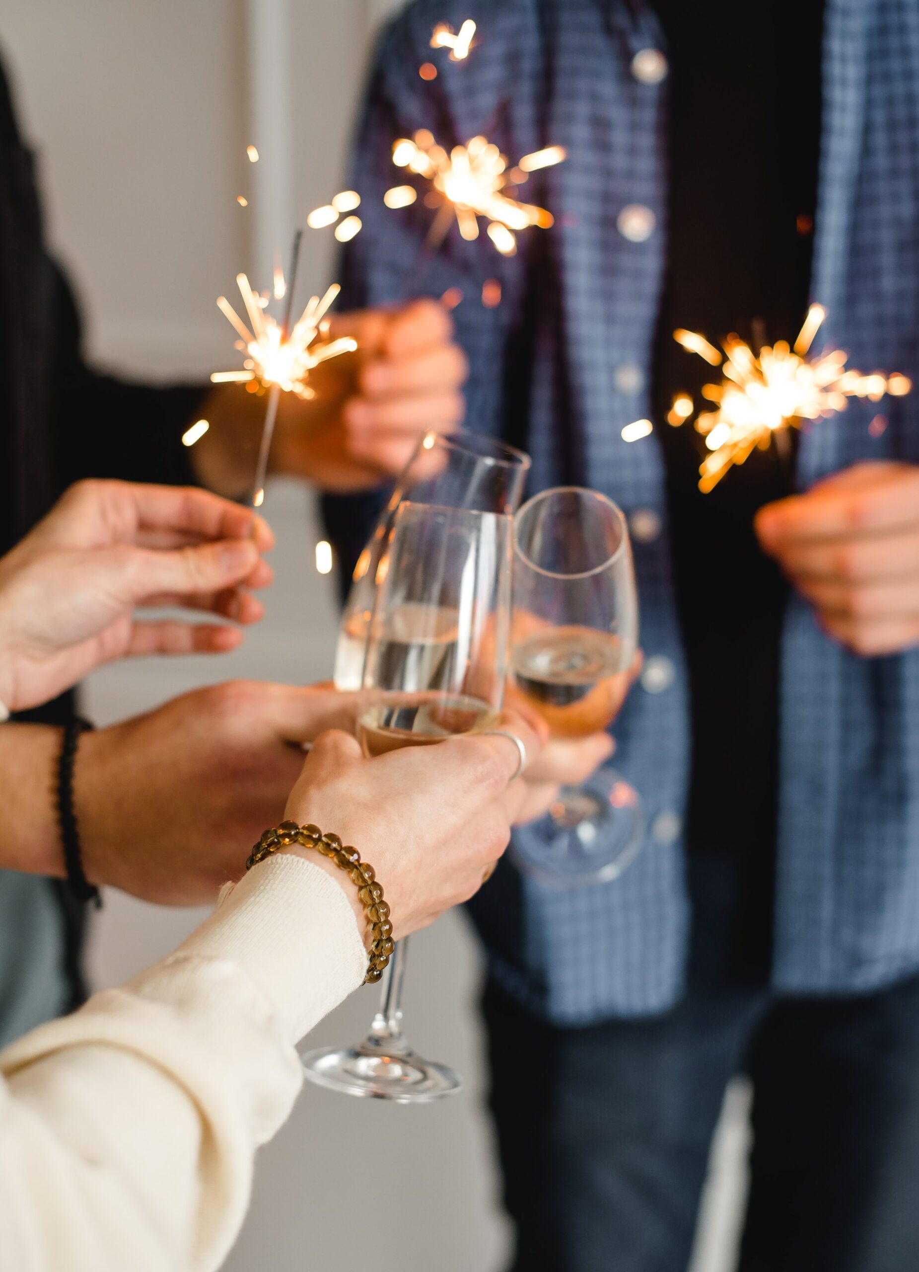 People clinking glasses and holding sparklers. The image captures a festive atmosphere with participants wearing casual clothing, including a blue plaid shirt. The focus is on the hands and sparklers, creating a lively, celebratory scene.