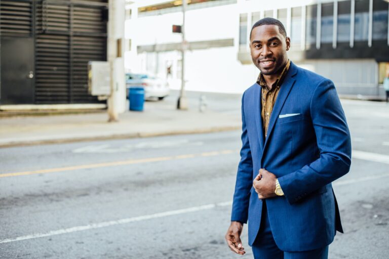 A man in a blue suit with a gold watch stands smiling on a city street. He holds his jacket with one hand and the background shows a building, a sidewalk, and a parked white car.