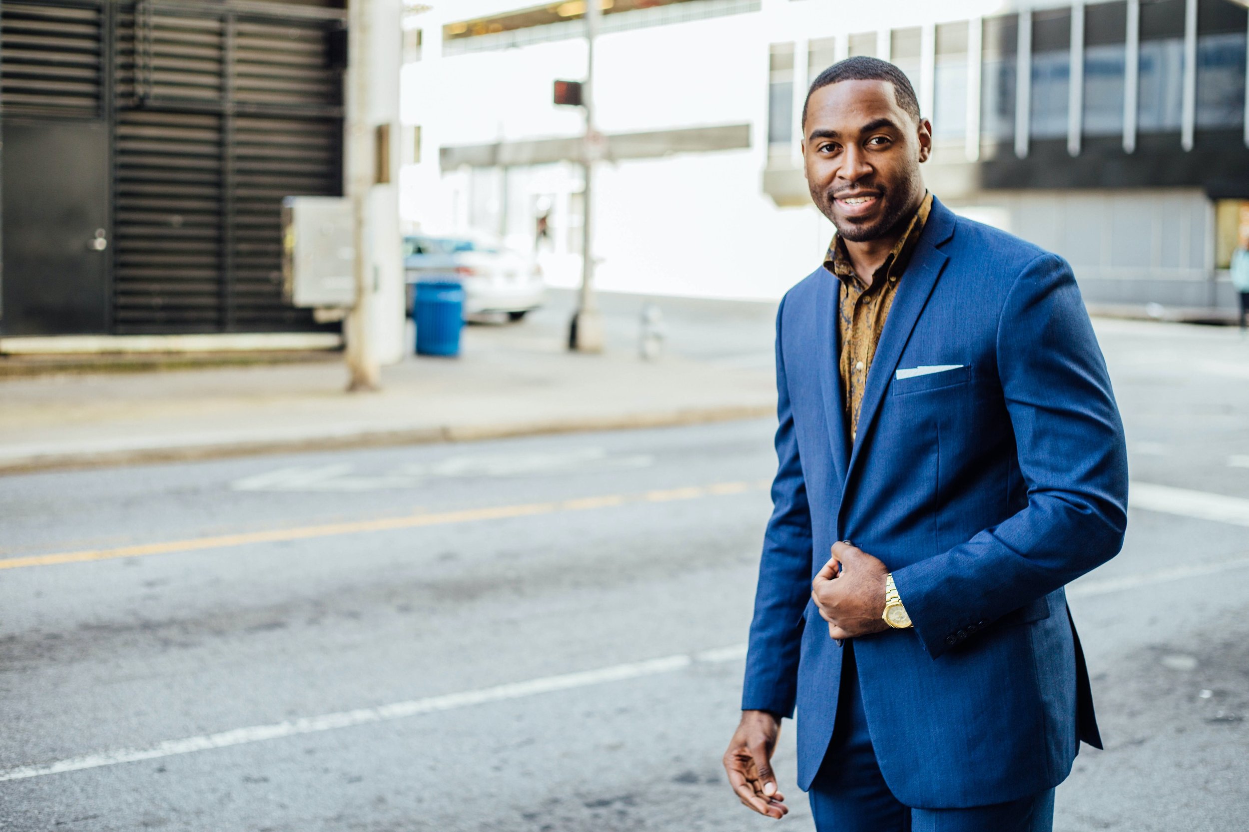 A man in a blue suit with a gold watch stands smiling on a city street. He holds his jacket with one hand and the background shows a building, a sidewalk, and a parked white car.
