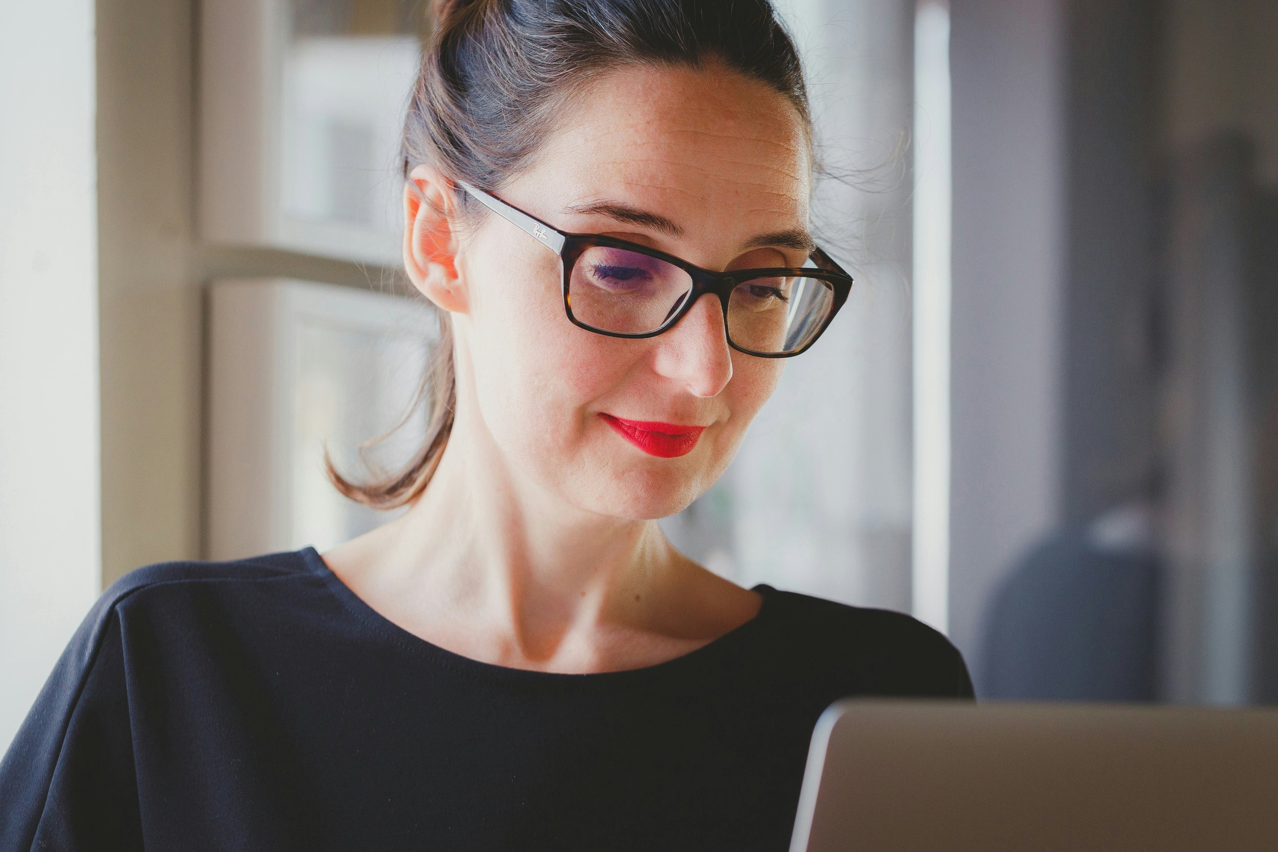 A person with dark hair tied back, wearing glasses and a black top, is looking at a laptop screen with a slight smile. They are indoors, near a window with sunlight filtering in.