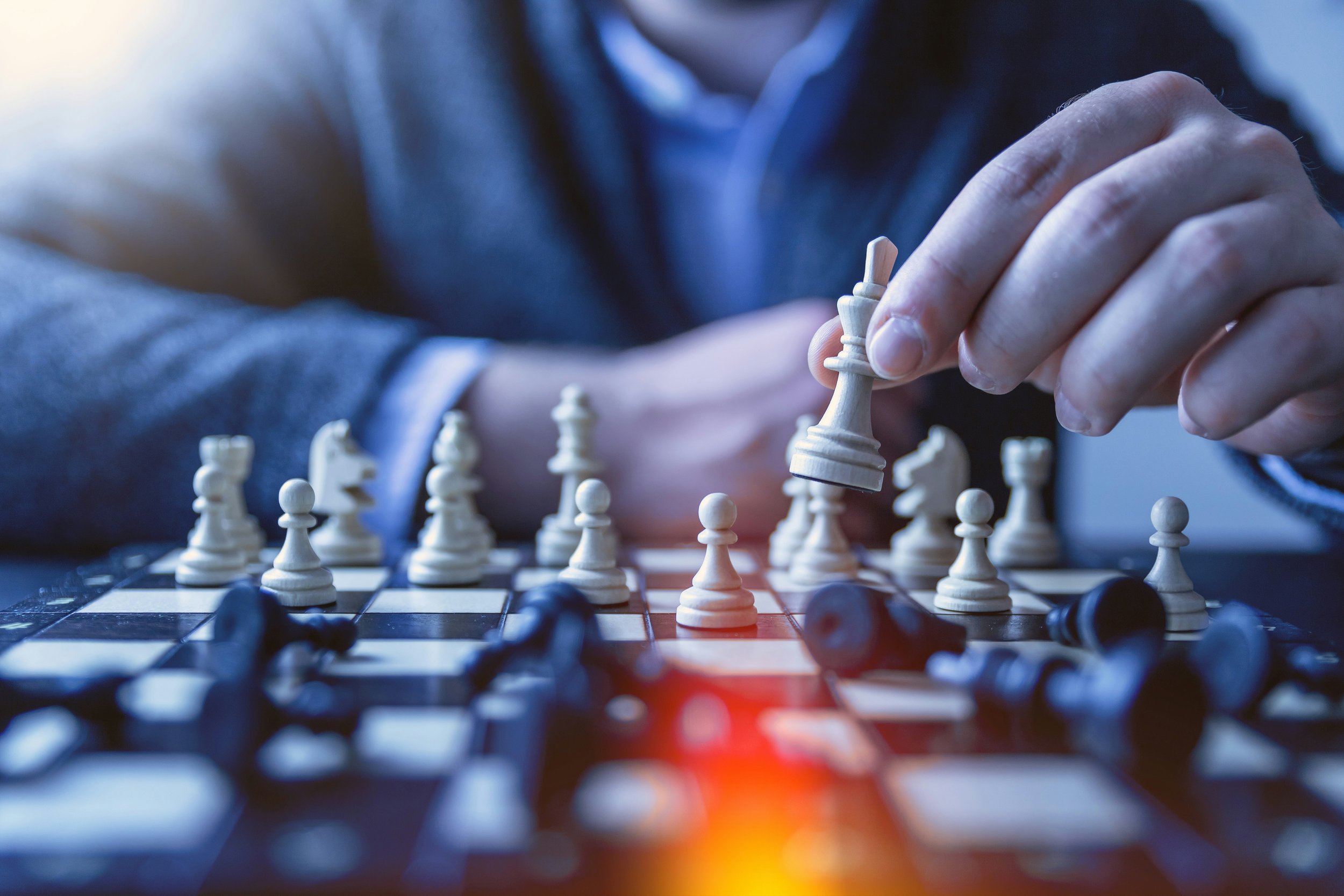 A person in a suit is holding a white king chess piece, hovering over a chessboard. Various pieces are positioned on and around the board. The lighting is warm, creating a focused and strategic atmosphere.