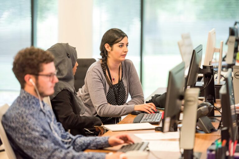 Three people focused on computers at a long desk in a bright office. The first person, wearing headphones, is in the foreground. The other two, one wearing a headscarf, are collaborating on a task. Large windows with blinds are in the background.
