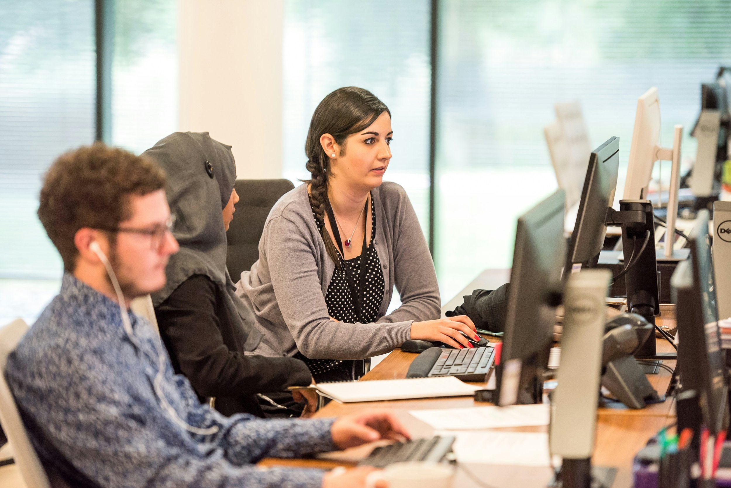 Three people focused on computers at a long desk in a bright office. The first person, wearing headphones, is in the foreground. The other two, one wearing a headscarf, are collaborating on a task. Large windows with blinds are in the background.