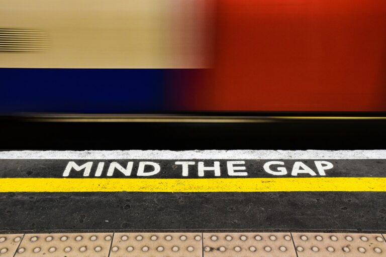 A blurred train speeds past an underground platform. The phrase "MIND THE GAP" is prominently painted in white on the platform's edge, bordered by a yellow line. The platform surface has tactile paving for safety.