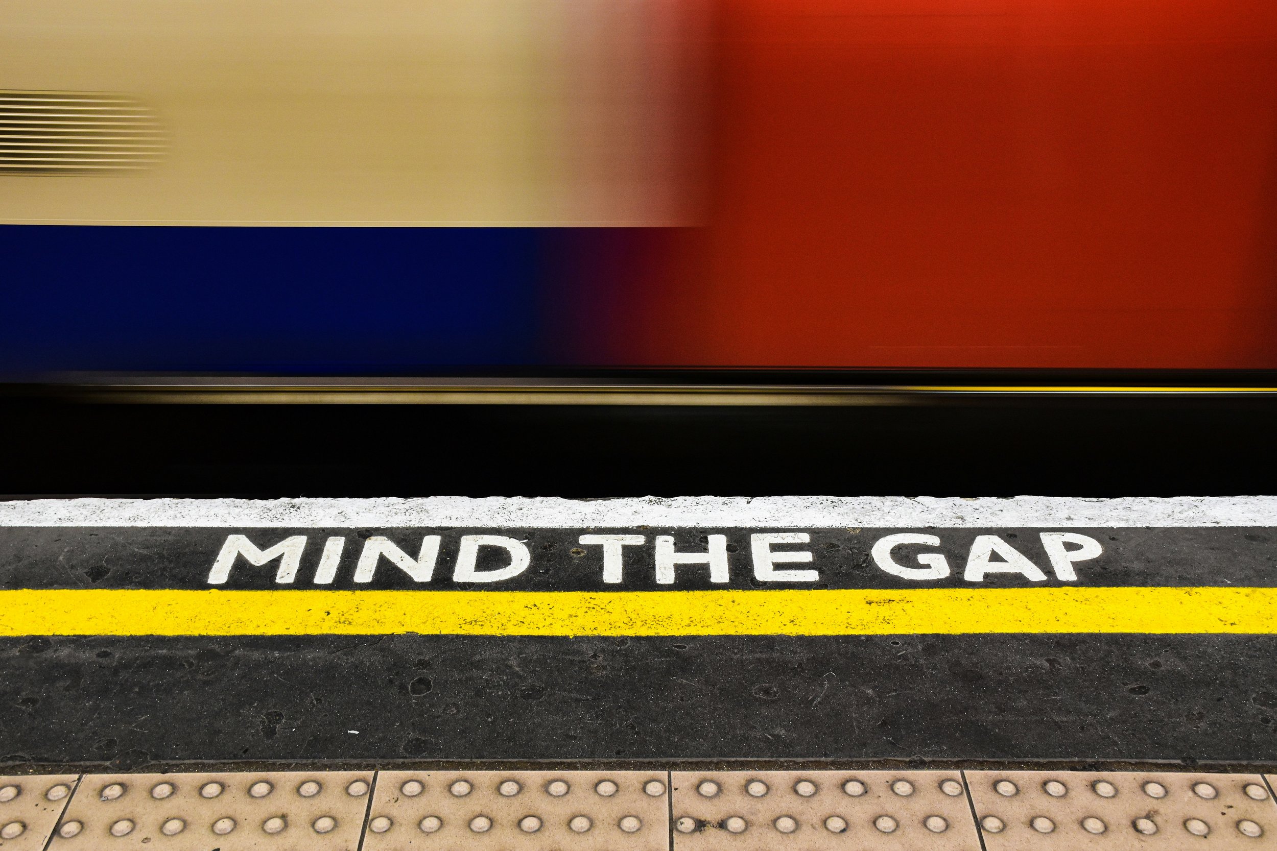 A blurred train speeds past an underground platform. The phrase "MIND THE GAP" is prominently painted in white on the platform's edge, bordered by a yellow line. The platform surface has tactile paving for safety.