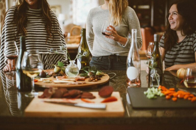 Four people gather around a kitchen island with wine glasses and appetizers. There are bottles and trays with various foods, including veggies and cured meats. The scene is warm and casual, with the group laughing and enjoying their time together.