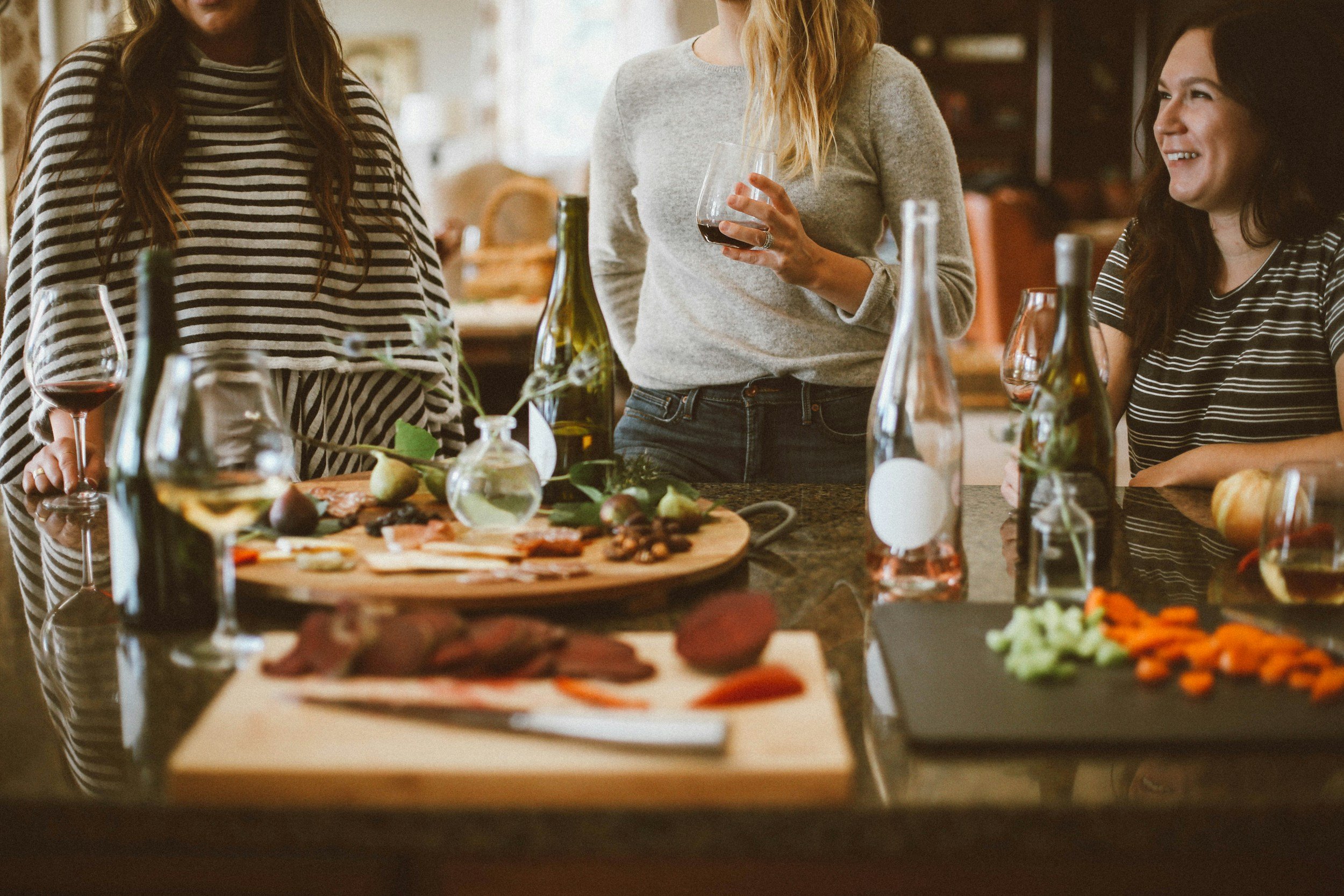 Four people gather around a kitchen island with wine glasses and appetizers. There are bottles and trays with various foods, including veggies and cured meats. The scene is warm and casual, with the group laughing and enjoying their time together.
