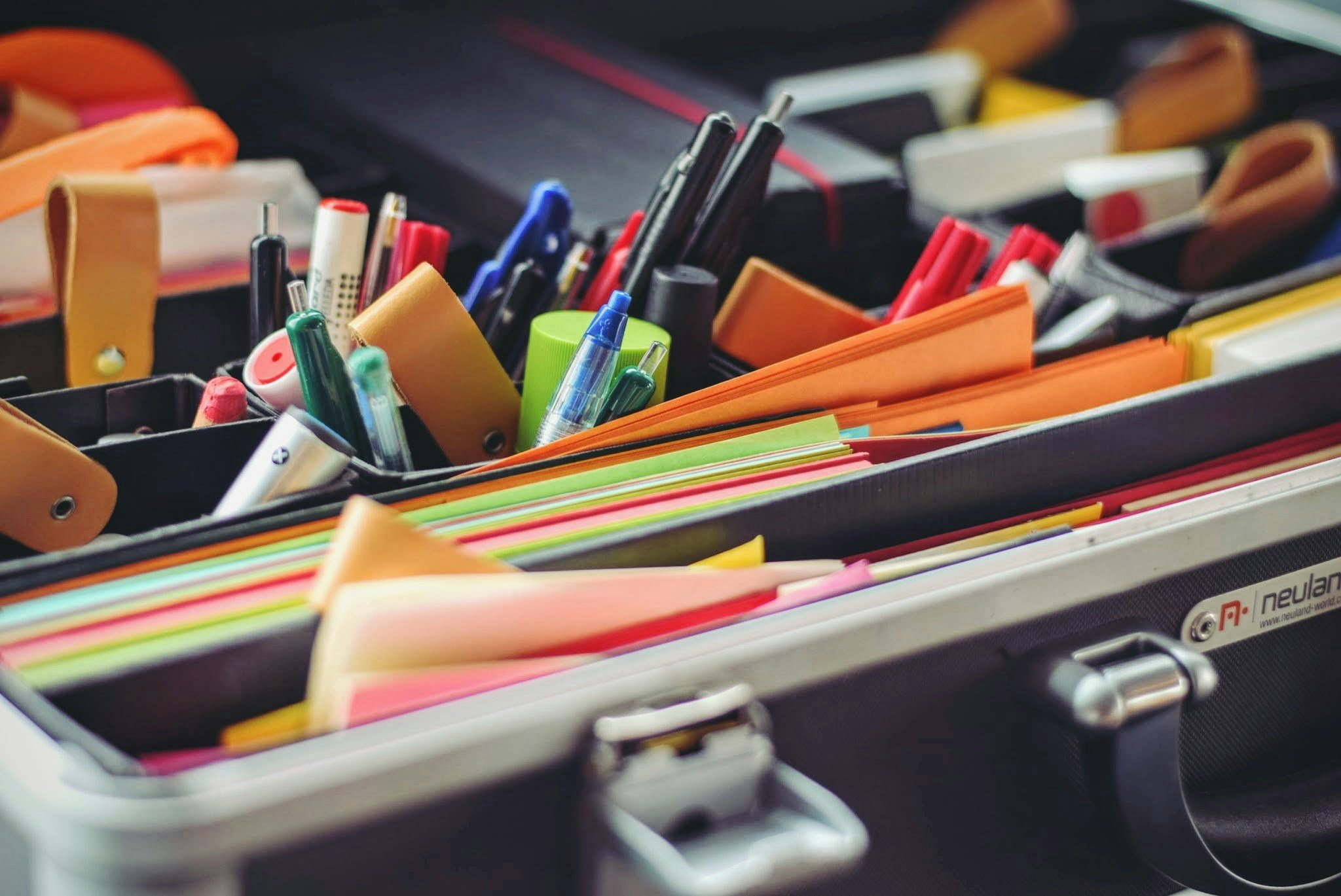 An open briefcase filled with various office supplies, including pens, highlighters, scissors, and colorful paper folders. The items are neatly organized in compartments, with some pens visible in pen holders.