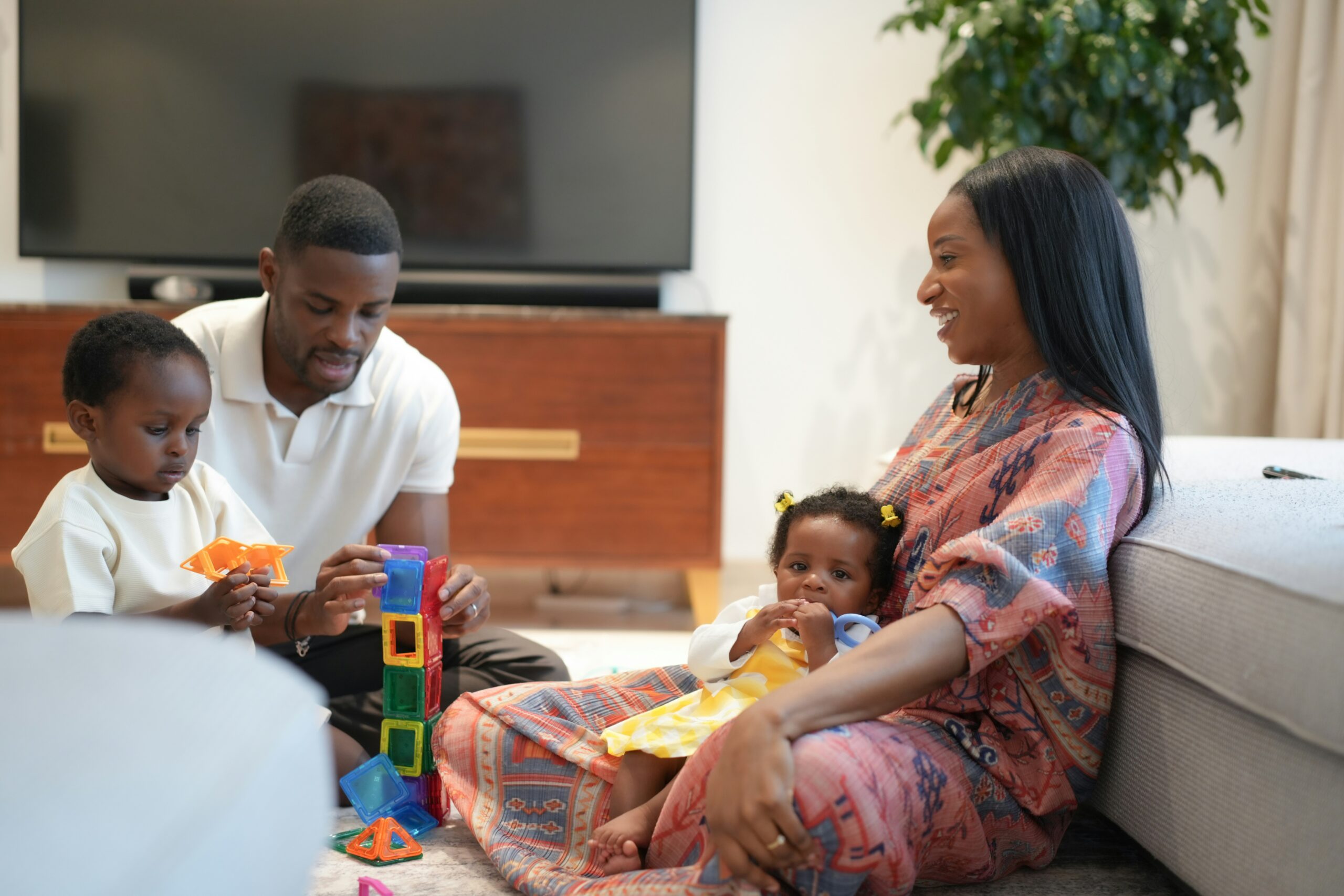 A family of four sits on the living room floor. A man helps a young child build with colorful blocks, while a woman smiles, holding a baby on her lap. A TV and plant are in the background.