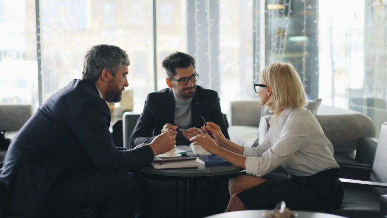 Three business professionals, two men and one woman, sit at a round table in an office, engaged in discussion. They hold pens and papers, appearing focused and collaborative in a modern, well-lit setting.