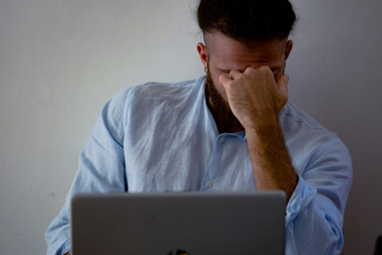 A man in a light blue shirt sits in front of a laptop, covering his face with his hand, appearing stressed or frustrated.