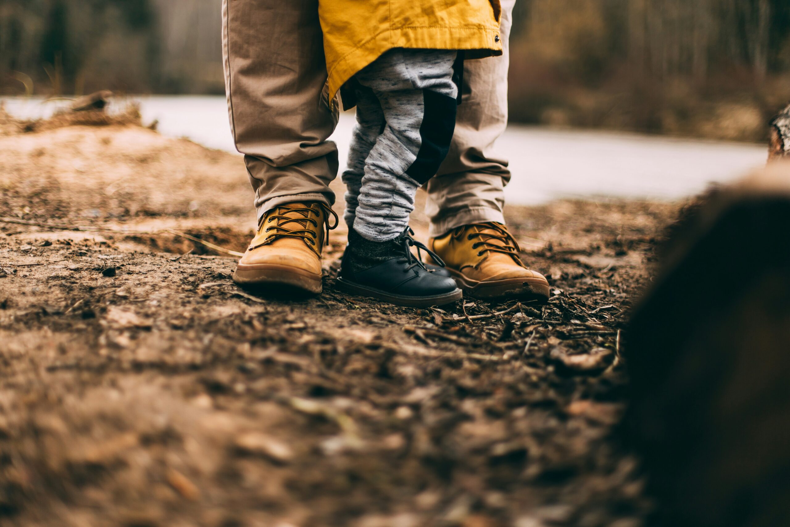 A child standing close to an adult outdoors. The adult wears tan boots and khaki pants, while the child wears black shoes, gray pants, and a yellow jacket. The ground is dirt with blurred trees and a river in the background.