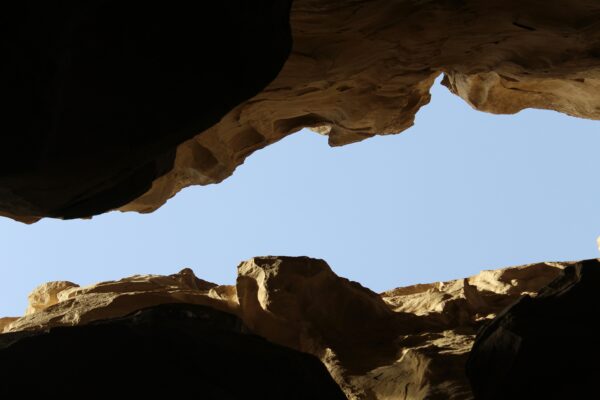 View from inside a rocky cave looking up at a bright blue sky, with rugged, sunlit stone walls framing the opening above.