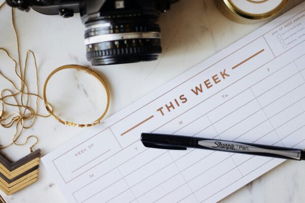 A weekly planner with “This Week” at the top sits on a desk with a Sharpie pen, a camera, a gold bracelet, and a necklace nearby. The planner has empty days and a space to write the week’s dates.