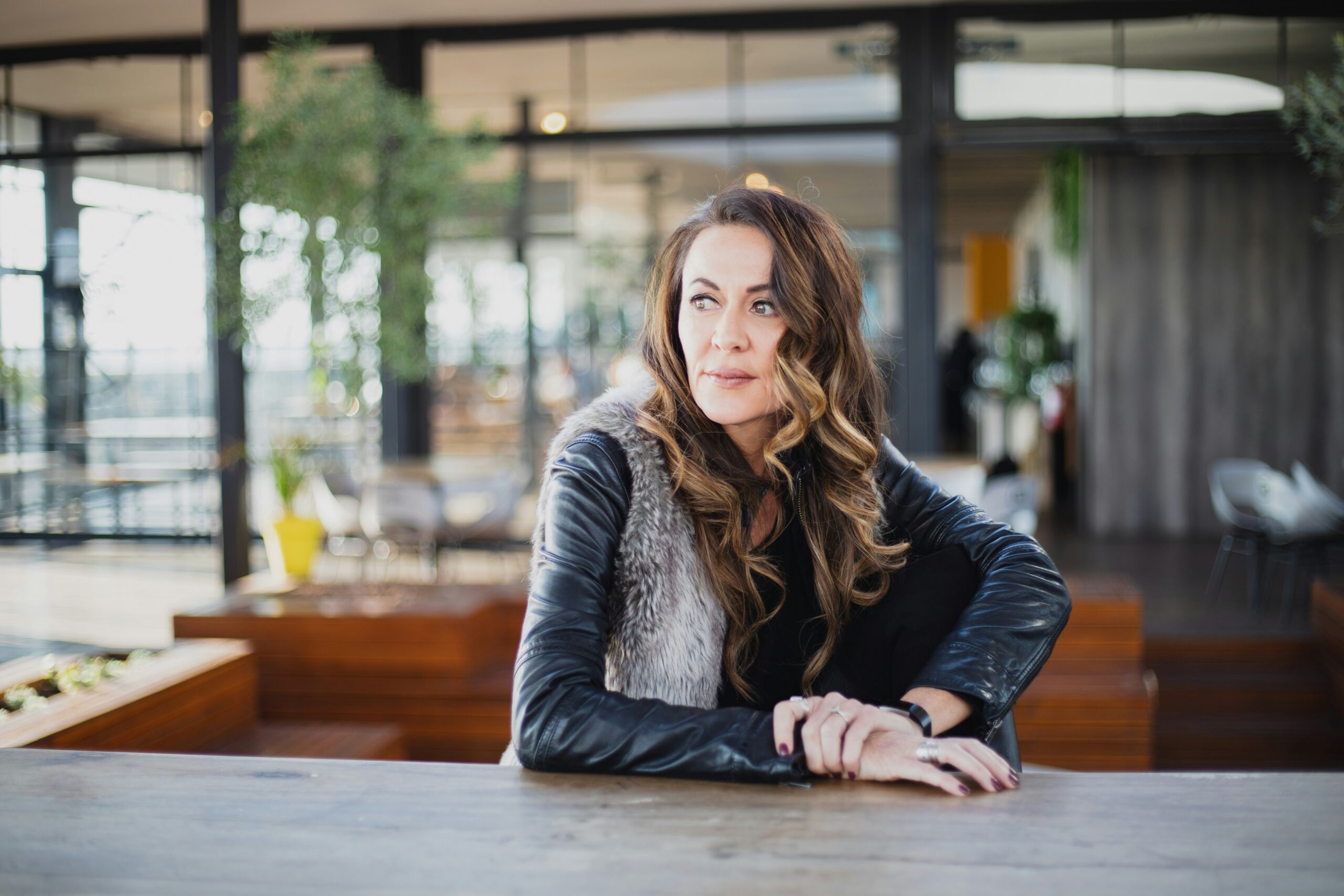 A woman with long, wavy hair wearing a leather jacket with a fur collar sits at a wooden table indoors, looking thoughtfully to the side in a modern, well-lit space with plants and glass walls.