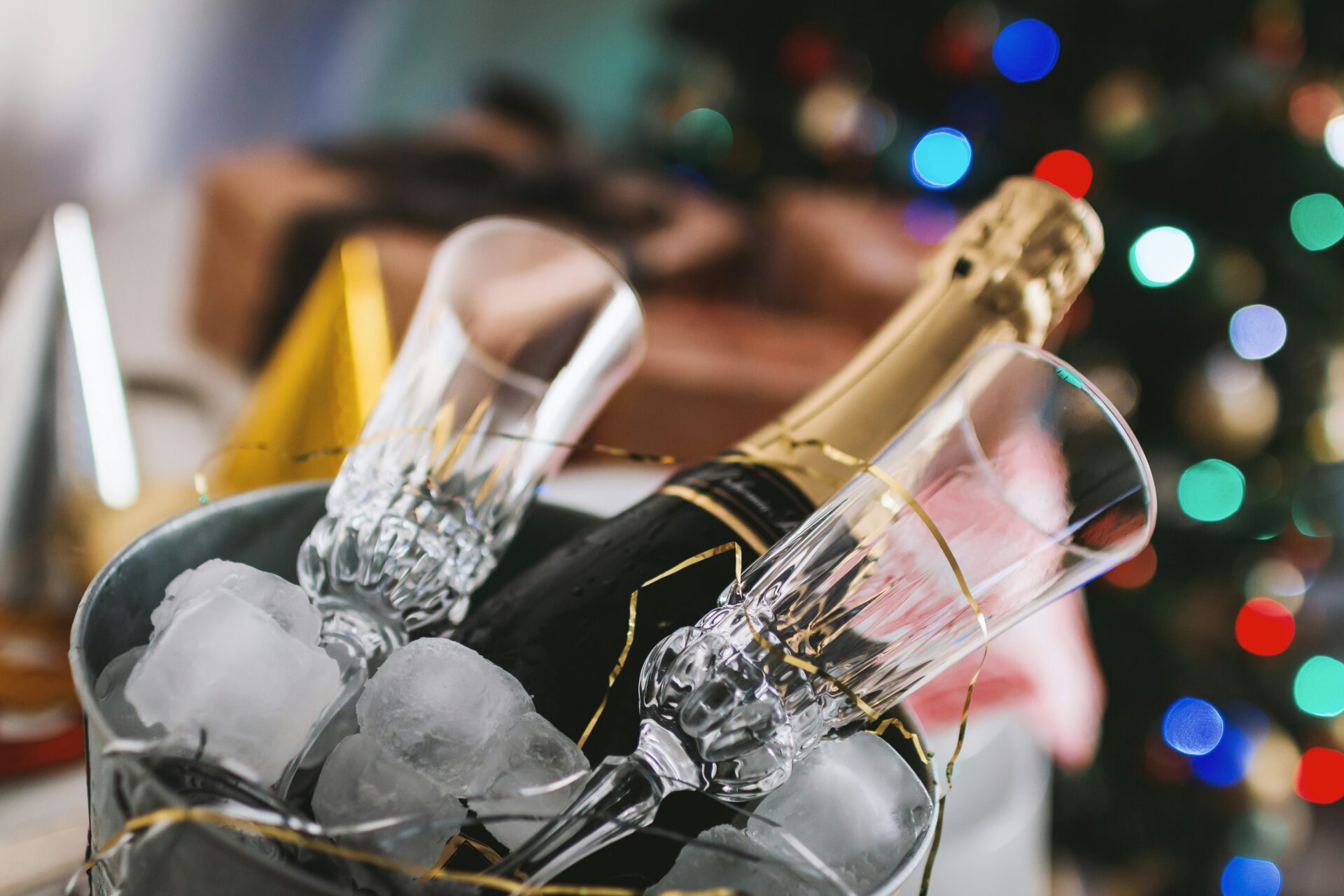 Two empty champagne flutes and a bottle of champagne rest in an ice bucket. In the background, blurred colored lights and wrapped presents suggest a festive celebration, possibly during the holidays.