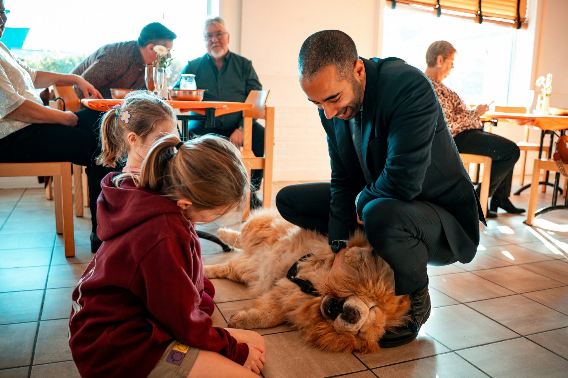 A man in a suit pets a large dog lying on the floor, while two young girls watch. Several adults sit at tables in the background in a brightly lit room.