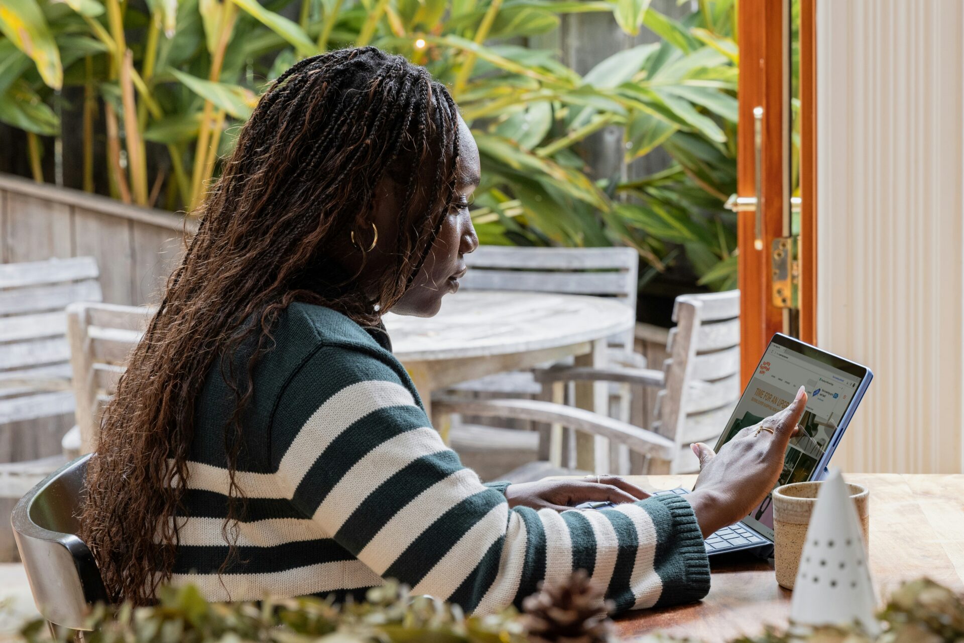 A person with long braided hair sits at a wooden table, using a tablet or laptop with a touch screen. Green plants and outdoor furniture are visible through open doors in the background.
